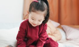 A young child in a red outfit is sitting and looking down, surrounded by cozy pillows in a softly lit room.