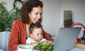 A woman, smiling, types on a laptop with a baby on her lap in a cozy room featuring a plant, toy stack, and a cup nearby.