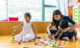 Children and an adult play with colorful wooden blocks on a wooden floor. One child is stacking blocks, while the adult holds another child, helping with the blocks. A bright room with shelves surrounds them.