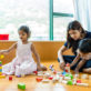 Children and an adult play with colorful wooden blocks on a wooden floor. One child is stacking blocks, while the adult holds another child, helping with the blocks. A bright room with shelves surrounds them.
