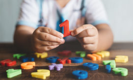 Child's hands holding a red number one, surrounded by colorful magnetic numbers scattered on a wooden table, suggesting a playful learning environment.