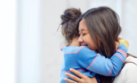 A young woman and a child cuddle affectionately, wearing brightly colored clothing, in a bright room with blurred white walls in the background.