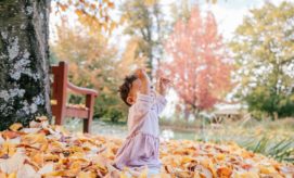 A toddler in a pink dress sits among fallen leaves, reaching upward under a large tree. Nearby, a wooden bench and vibrant autumn foliage enhance the peaceful park setting.