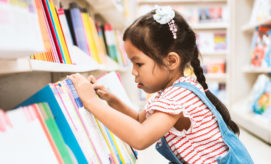 A young child wearing a striped shirt and overalls is selecting a book from a colorful shelf in a bookstore or library. Shelves filled with books surround her.