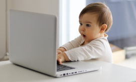 A baby interacts with a laptop, pressing keys while sitting at a white table. The setting is a bright room with natural light streaming in from a window.