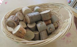 A wicker basket holds several small, cylindrical wood pieces. The basket is on a table, and the wood appears naturally cut, with visible bark and smooth ends.