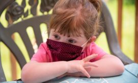 A young child wearing a patterned face mask rests their chin on folded arms at a table, sitting on an ornate chair, with a blurred green background.
