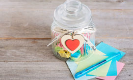A glass jar filled with colorful paper stars and a wooden heart tag labeled “Handmade” rests on a wooden surface beside colorful paper squares and a blue pen.