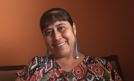 A person with long earrings and a patterned shirt is smiling while seated in front of a warm, brown wall.