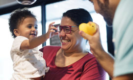 Child reaching towards a smiling adult's rubber duck toy, with another adult holding the child in a brightly lit indoor setting.