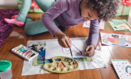 A child paints on paper with a brush from a palette, surrounded by art supplies and colorful drawings, while lying on a wooden floor with a purple rug nearby.