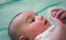A baby lies on a soft, green blanket, looking intently at their own hands, wearing a light-colored outfit with minimal patterns.