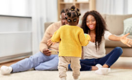 A toddler in a yellow sweater walks towards two smiling adults sitting on the floor with open arms, in a cozy living room setting.