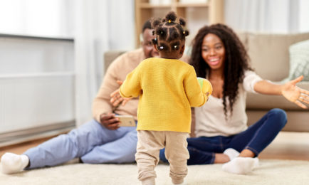A toddler in a yellow sweater walks towards two smiling adults sitting on the floor with open arms, in a cozy living room setting.