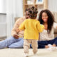 A toddler in a yellow sweater walks towards two smiling adults sitting on the floor with open arms, in a cozy living room setting.