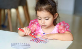 A young child, wearing a pink dress, colors with a red marker on paper at a table in a softly lit room, focused intently on the drawing.