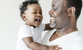 A smiling man holds a laughing baby, both wearing white shirts, in a bright, minimalistic space.