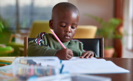Child drawing with a colored pencil, concentrating on their work, seated at a table in a bright room. Nearby are scattered crayons and paper, with plants in the background.