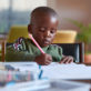 Child drawing with a colored pencil, concentrating on their work, seated at a table in a bright room. Nearby are scattered crayons and paper, with plants in the background.