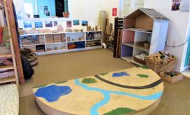 A wooden play area with painted roads, rivers, and trees lies on the carpet. Shelves filled with wooden toys and blocks line the back wall in a brightly lit room.