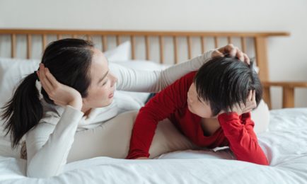 A woman and child lie on a bed, facing each other, in a relaxed, intimate conversation. The bed has white sheets and a wooden headboard, creating a cozy atmosphere.