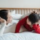 A woman and child lie on a bed, facing each other, in a relaxed, intimate conversation. The bed has white sheets and a wooden headboard, creating a cozy atmosphere.