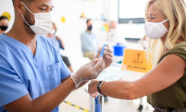 A healthcare worker in blue scrubs prepares a syringe for a patient wearing a mask. The setting appears to be a vaccination clinic with people seated in the background.
