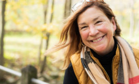 A woman smiles warmly, standing outdoors. Her hair is gently blown by the wind. She wears a brown vest and scarf, surrounded by blurred autumnal trees in soft focus.