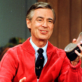 A person wearing a red cardigan and tie holds a shoe, smiling warmly. The setting appears indoors with blurred greenery in the background.