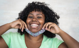 A person with curly hair smiles while lowering a floral face mask, wearing a light green shirt against a plain, light gray background.