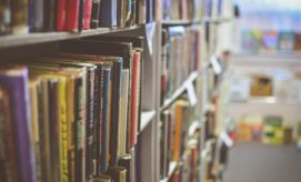 Books line shelves, tightly packed, in a dimly lit library aisle. The blurred background reveals more books and soft light filtering through windows, suggesting a peaceful, studious environment.