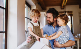 A man interacts with two children, one standing and one in his arms, in a bright, cozy room with large windows, creating a warm family scene.