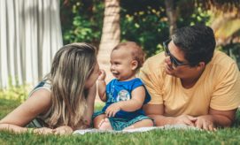 A baby in a blue outfit sits in a grassy park, smiling and interacting with two adults lying beside them. Trees and a curtain in the background provide a shaded setting.