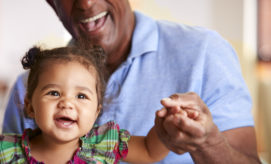 A toddler, smiling and wearing a colorful plaid dress, is held and supported by an adult wearing a blue shirt, both appearing joyful in a warmly lit indoor setting.