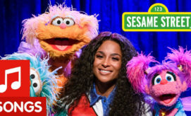 A person smiles while surrounded by colorful Sesame Street puppets under blue stage lights. The Sesame Street sign is visible above. Text: "SONGS".