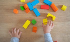 Children's hands play with colorful wooden blocks on a light wooden table, organizing them into various shapes and structures.