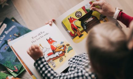 Child and adult reading a picture book about a fox, seated on a cozy wooden floor. The page shows the fox searching for socks, with various books scattered nearby. Text reads, “Poor old Fox has lost his socks.”