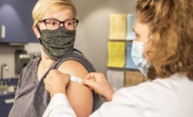 A healthcare worker applies a bandage to a patient's upper arm in a medical clinic, both wearing face masks, with cabinets and informational posters in the background.