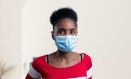 A person wearing a blue surgical mask stands in a room with light-colored walls, wearing a red and white striped shirt, conveying a calm demeanor.