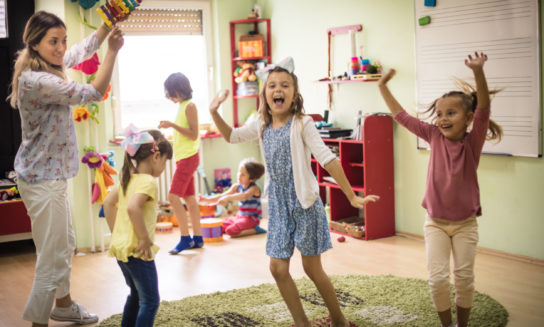 Children energetically dance in a classroom with brightly decorated walls, guided by a teacher holding up a colorful piñata. Toys and shelves line the background, creating a playful atmosphere.