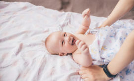 A smiling baby lies on a patterned blanket, raising its arms while an adult's hands gently adjust the baby’s wrap, creating a warm, nurturing scene.