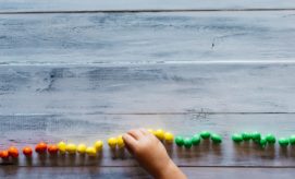 A child's hand arranges colorful candies in a line by hue on a rustic wooden table, transitioning from red to green with a natural light setting.