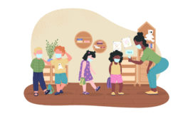 Children wearing masks stand in line as a teacher checks a student's temperature with a digital thermometer in a classroom. Shelves and wall decorations surround them.