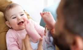 A smiling baby wearing a pink shirt lies on a brown surface, playfully interacting with an adult who is holding the baby's feet, in a cozy indoor setting.