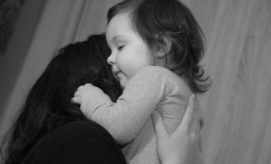 A child, eyes closed, is being gently held in someone's arms, creating a tender embrace. The setting appears to be indoors, with soft lighting illuminating their profiles.