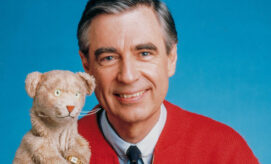 A person smiles warmly, holding a tan puppet of a cat. They wear a red cardigan against a plain blue background. The puppet has a kind expression with wide eyes.
