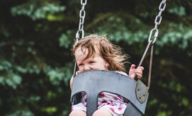 A young child smiles while sitting in a black swing, surrounded by blurred green trees in a playground setting.