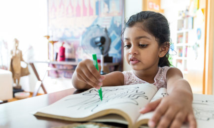 A young child colors a picture with a green crayon in a coloring book, seated at a table in a well-lit room with artwork and shelves in the background.