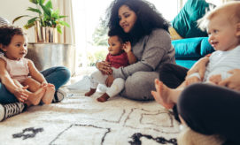 An adult sits on the floor, holding a baby in a cozy room. Three other babies sit nearby, with a plant and a turquoise couch in the background.