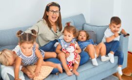A woman sits on a sofa, smiling, surrounded by five young children playing energetically. The children hold toys, and the setting is a bright, cozy living room.